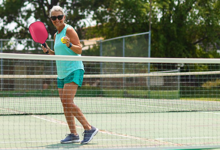 A woman smiles holding a pickleball paddle and ball while standing on a sports court about to serve to her partner