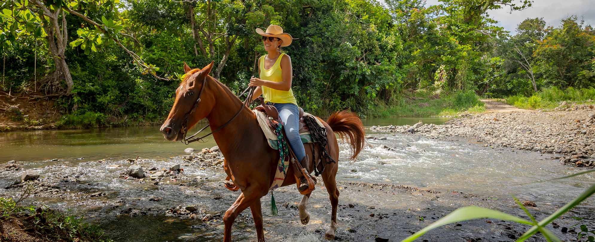 A woman wearing a yellow tank top and a wide-brimmed hat riding a horse through a rocky river. 