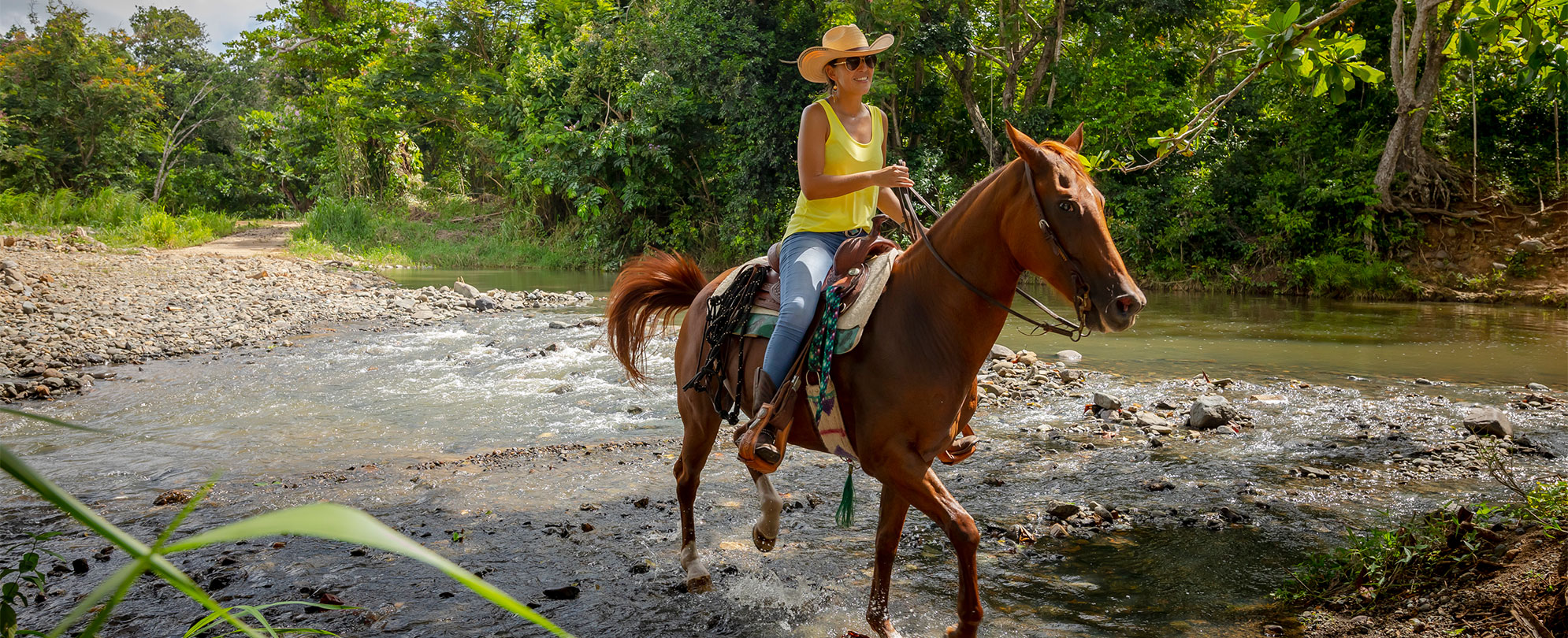 Woman wearing a yellow tank top and a wide-brimmed hat riding a horse through a rocky river. 