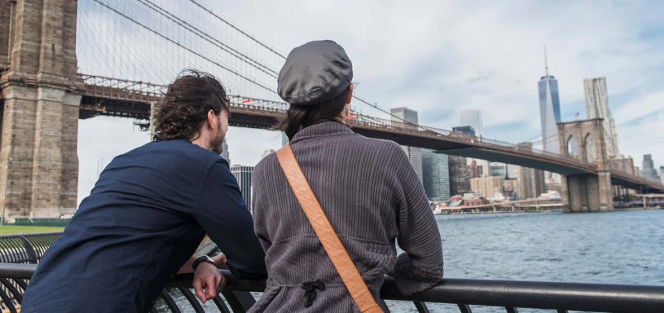 Man and woman leaned over railing, taking in the view of the New York City skyline and Brooklyn Bridge.