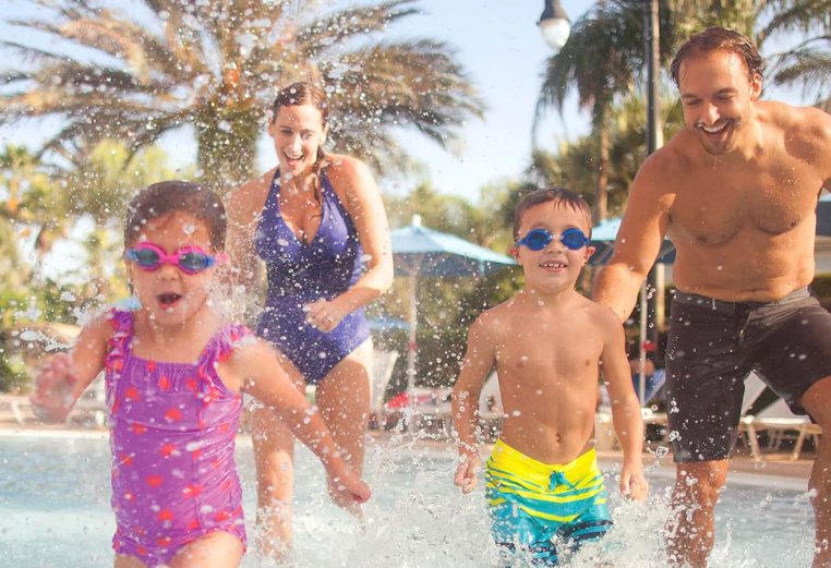 Parents with two young kids wearing goggles run through a splashpad at a timeshare resort.