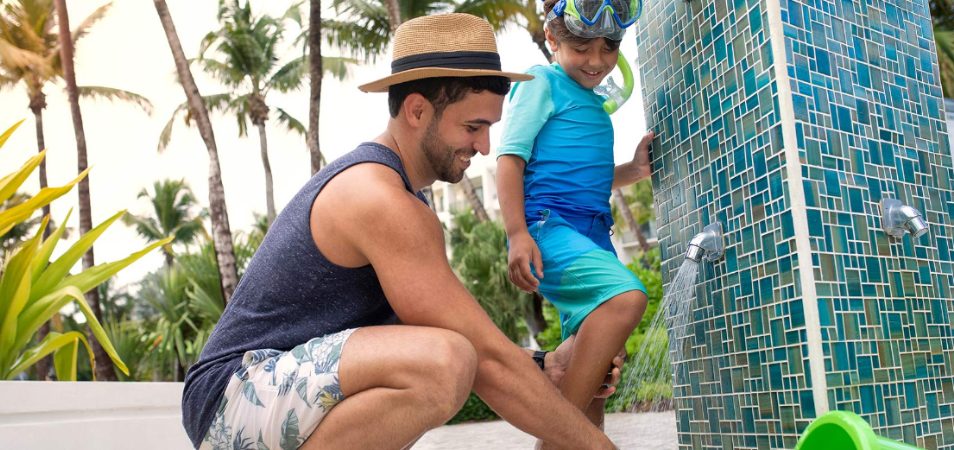 A dad helping his son wash sand off his feet in an outdoor shower at a Margaritaville Vacation Club resort.