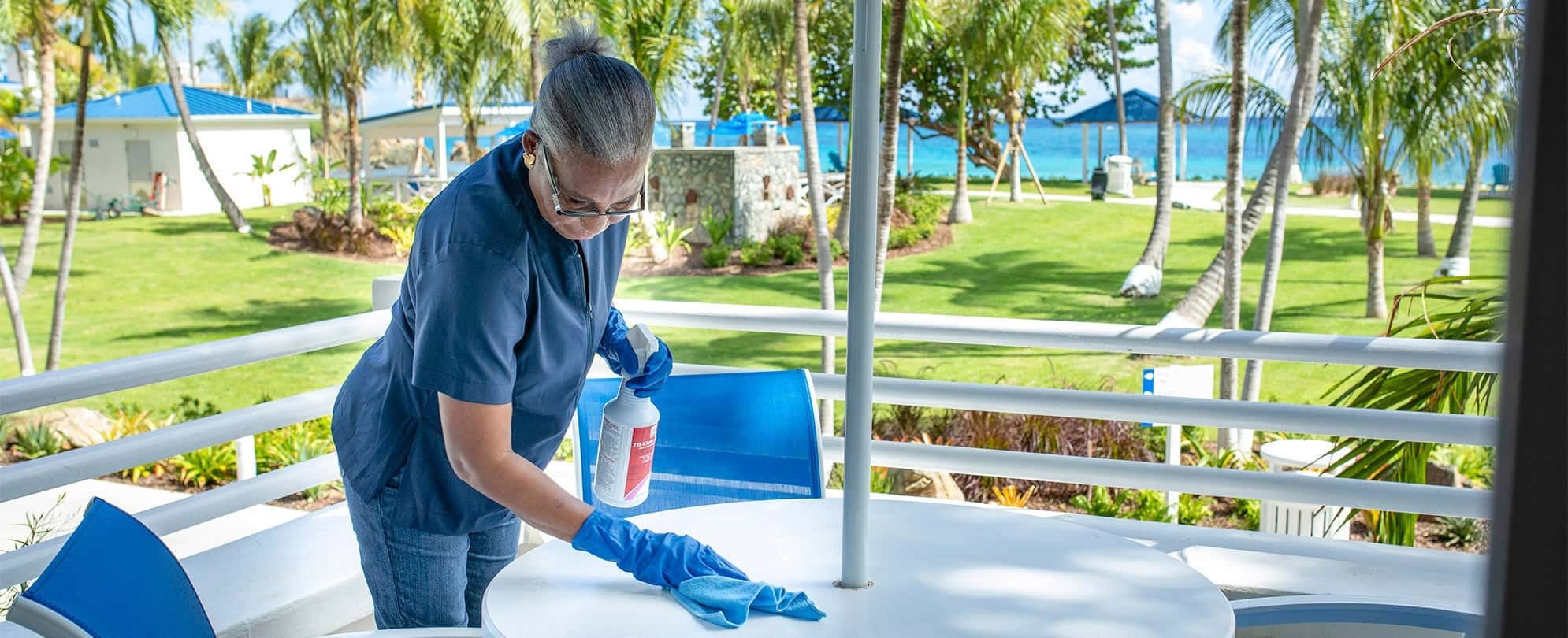 Female employee wearing a blue medical mask and gloves wipes an outdoor table at a Margaritaville Vacation Club resort. 