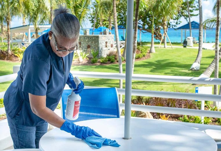 Female employee wearing a blue medical mask and gloves wipes an outdoor table at a Margaritaville Vacation Club resort. 