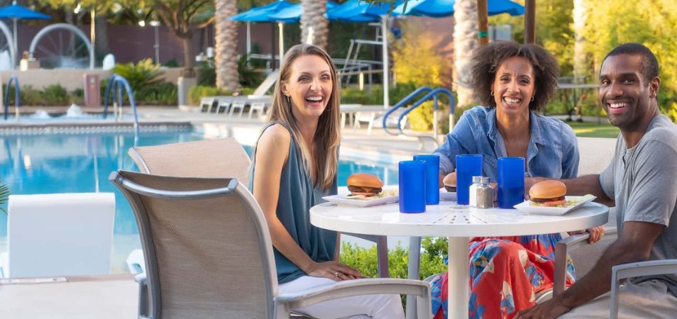 Three smiling adults enjoy burgers and drinks in blue cups at a poolside table of a Margaritaville Vacation Club resort.