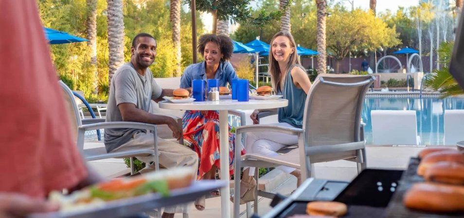 Three adults sitting at a poolside table enjoy grilled burgers at a Margaritaville Vacation Club resort.