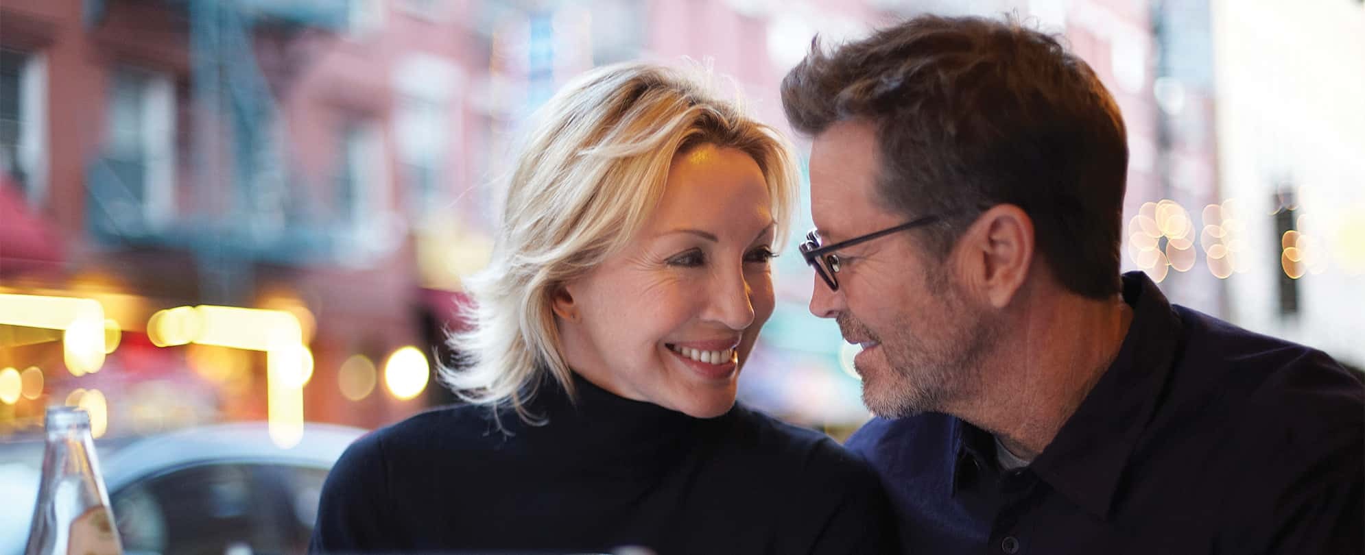 A man wearing glasses and woman in a black turtleneck shirt smile lovingly at each other.